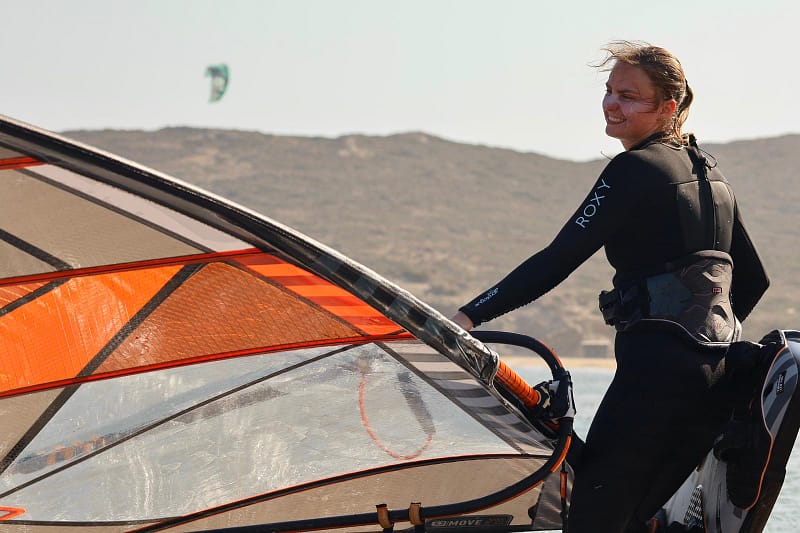 Uśmiechnięta kursanta idzie z deska windsurfingowa podczas wyjazdu Wavecamp na Rodos