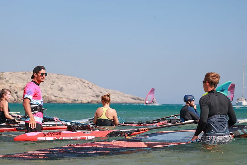 Grupa kursantów stoi w wodzie z instruktorem windsurfingu Wavecamp podczas zajęć na Rodos
